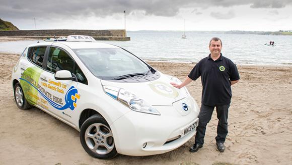 man standing next to a nissan leaf taxi on the beach